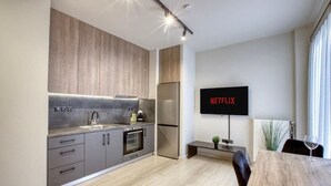 Uma cozinha moderna com armários cinzas, forno embutido e geladeira de aço inoxidável. Uma TV de tela plana na parede exibe "NETFLIX".