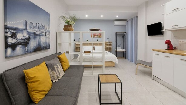 A modern hotel room with a grey sofa, a bed with a white headboard, a small wooden table, a kitchen area with white cabinets, and a large cityscape painting on the wall. - A little gem in the heart of Athens (Athina)