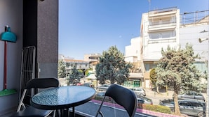 Un balcon avec une table ronde en verre et deux chaises, donnant sur une rue bordée de voitures garées et d’immeubles.