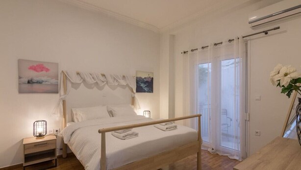 A bedroom with a wooden bed, white bedding, a nightstand with a lamp, and a view of the outdoors through a window with curtains.