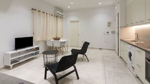 A modern apartment with a living area featuring a TV, a dining table with chairs, a kitchen with a washing machine, and a small seating area with chairs.