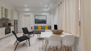 A modern kitchen with white cabinets, a microwave, and an oven. There is a dining area with a white table and chairs, and a sofa with yellow cushions.