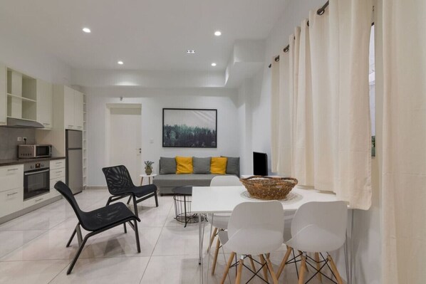 A modern kitchen with white cabinets, a microwave, and an oven. There is a dining area with a white table and chairs, and a sofa with yellow cushions. - Heaven apartment in the heart of Athens (Athina)