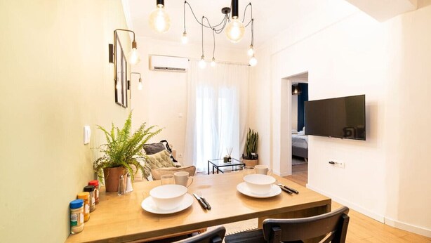 A modern dining area with a wooden table, chairs, and a chandelier. There is a TV mounted on the wall and a plant on the table.