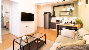 A modern living room with a sofa, a coffee table, and a TV. There is a kitchen in the background with a microwave, a sink, and a stove.