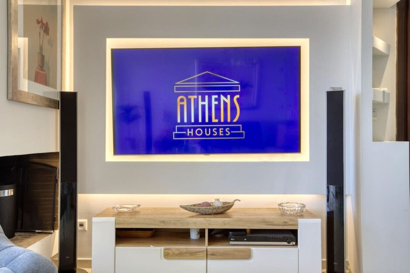 A modern living room with a mounted TV displaying 'ATHENS HOUSES' logo, wooden furniture, and a fireplace.