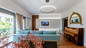 A modern living room with a sectional sofa, a wooden piano, a television, and a large window with curtains. - Luxury flat at archaeological museum (Athina)