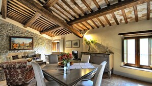 A rustic interior with exposed wooden beams, stone walls, a dining table with chairs, a sofa, and a window with natural light.