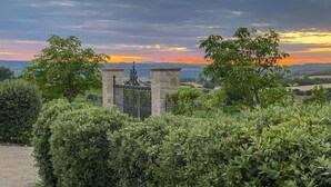 A gated entrance with stone pillars and a metal gate, surrounded by lush greenery and a scenic view of a sunset over a hilly landscape.