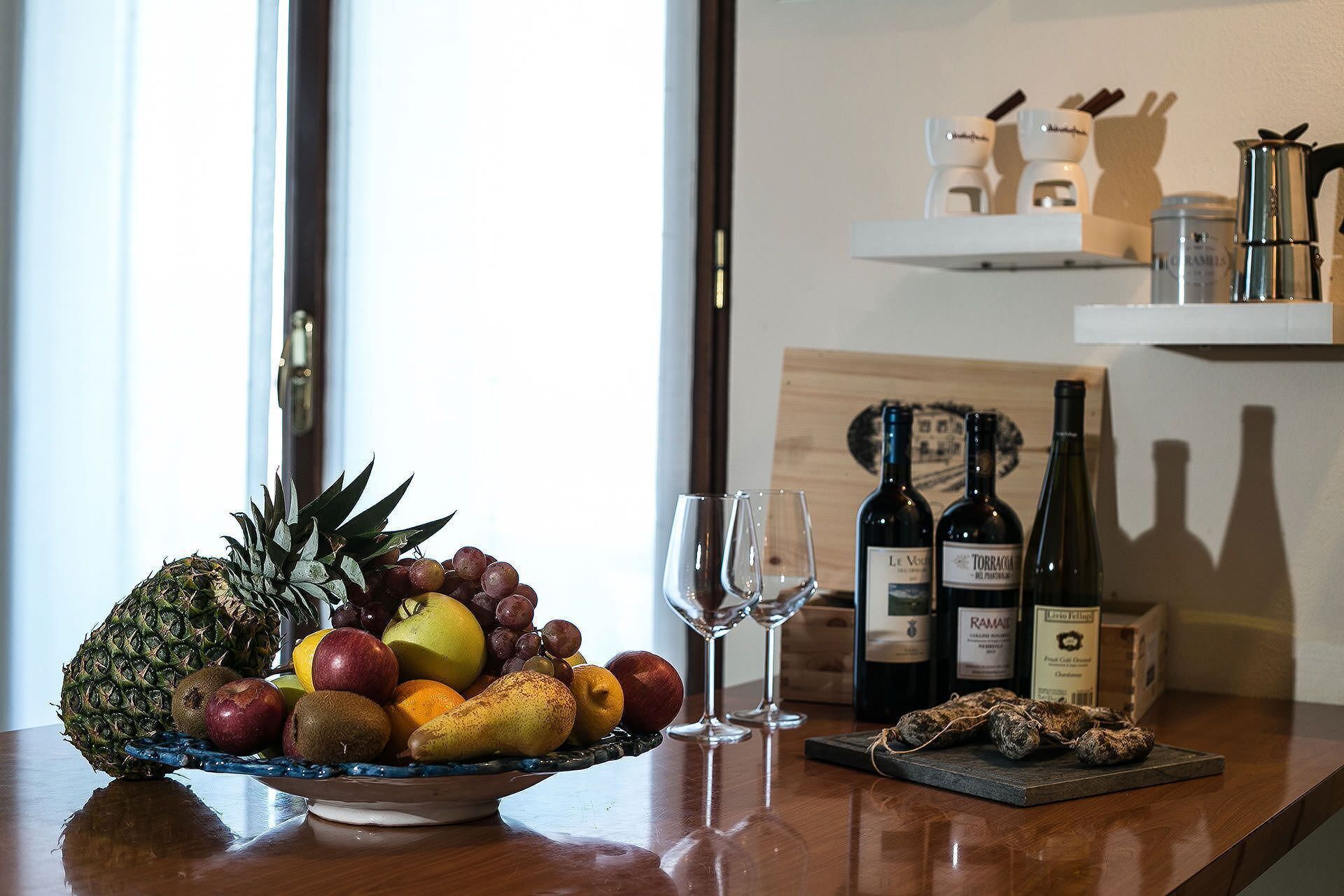 A fruit bowl with various fruits and two wine glasses on a wooden table.