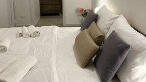 Un lit bien fait, avec du linge de lit blanc et des serviettes pliées, une table de chevet et une vue sur une salle de bain équipée d’un miroir et d’un lavabo.