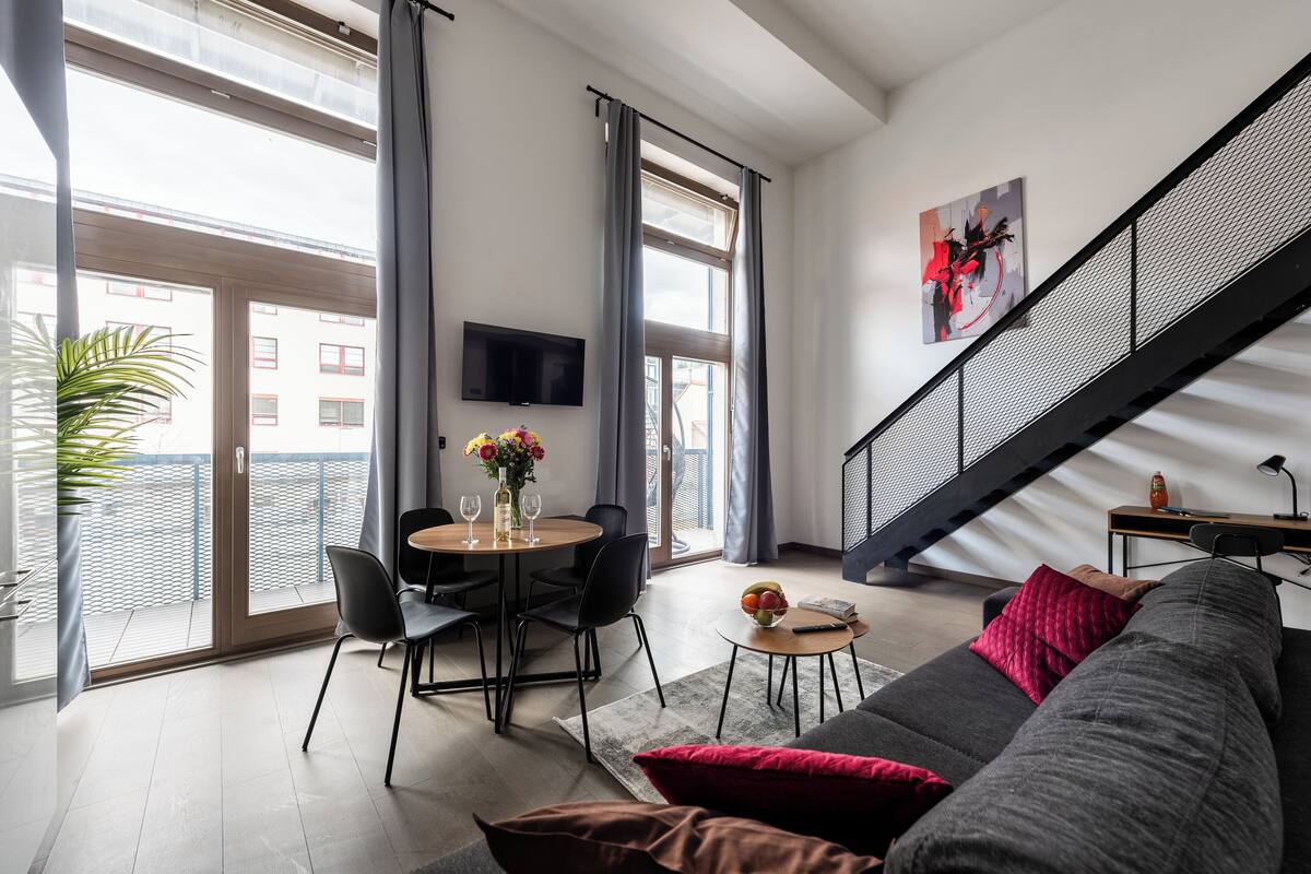 Een moderne woonkamer met een eethoek, grote ramen, een trap en een flatscreen-televisie.
