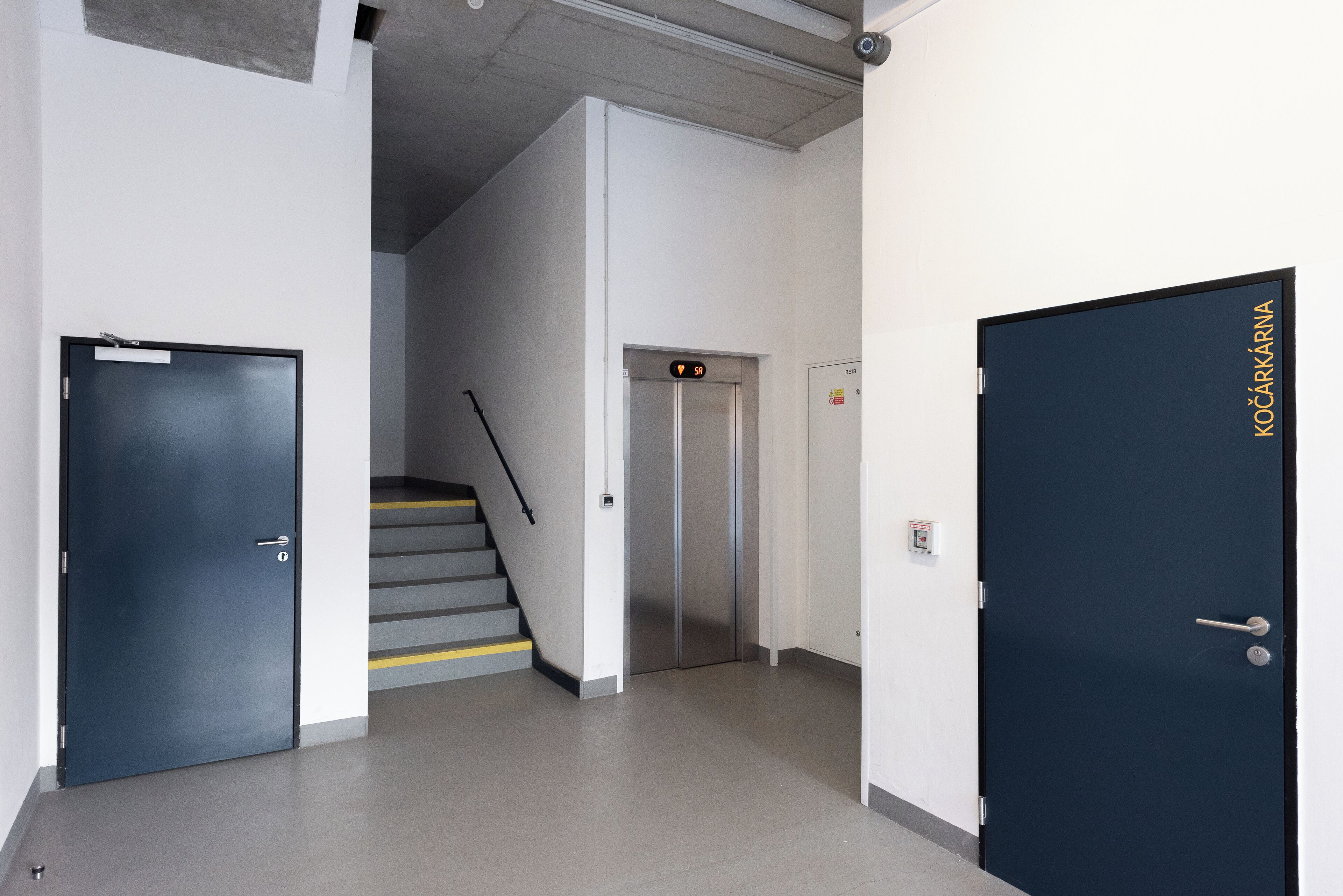 Un pasillo con dos puertas, una que dice "KOČARSKÁ" y otra cerrada. Se ve una escalera con una línea amarilla de seguridad al fondo.