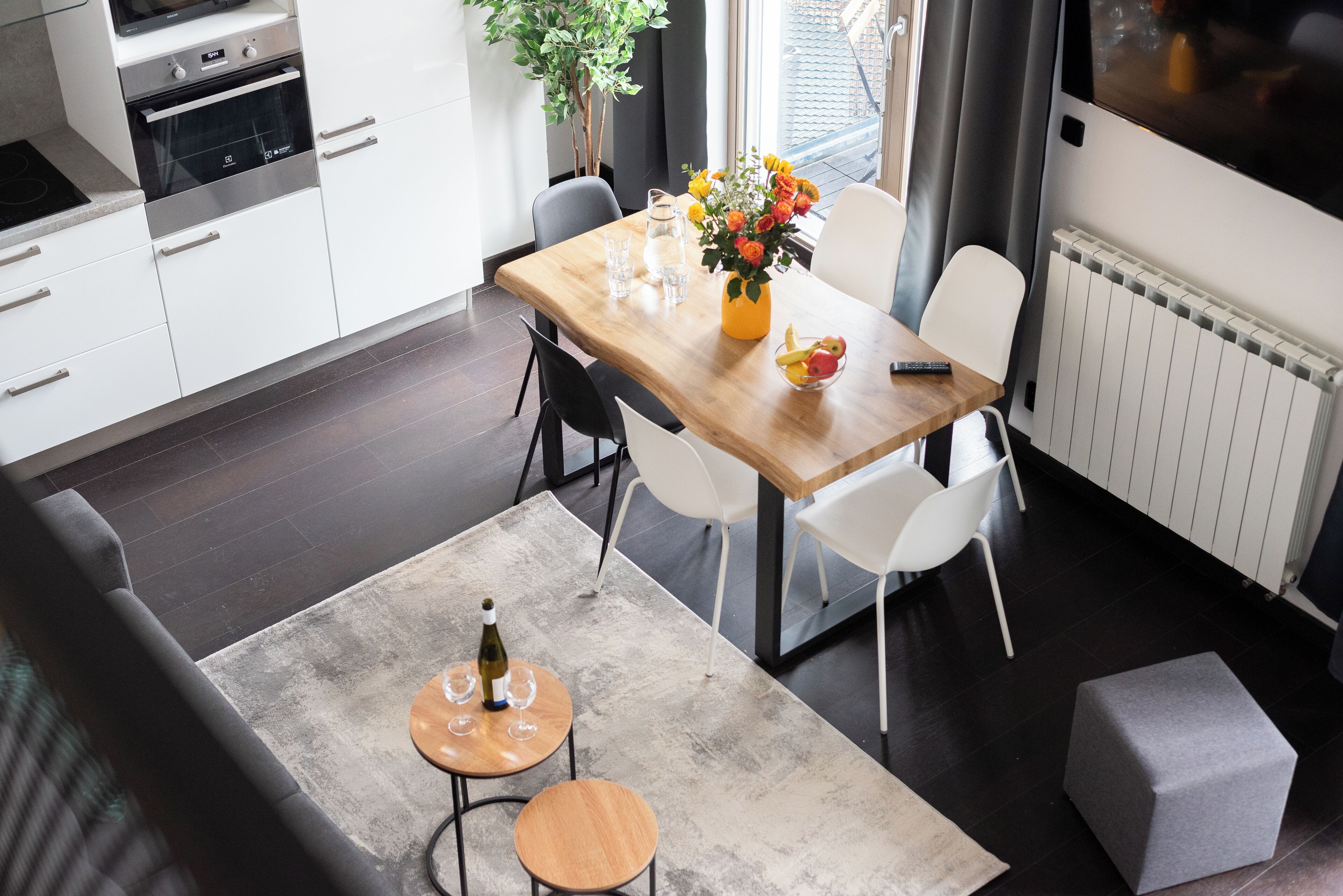 Une cuisine moderne avec des meubles blancs, une table à manger en bois et un petit coin salon agrémenté d’un pouf gris.