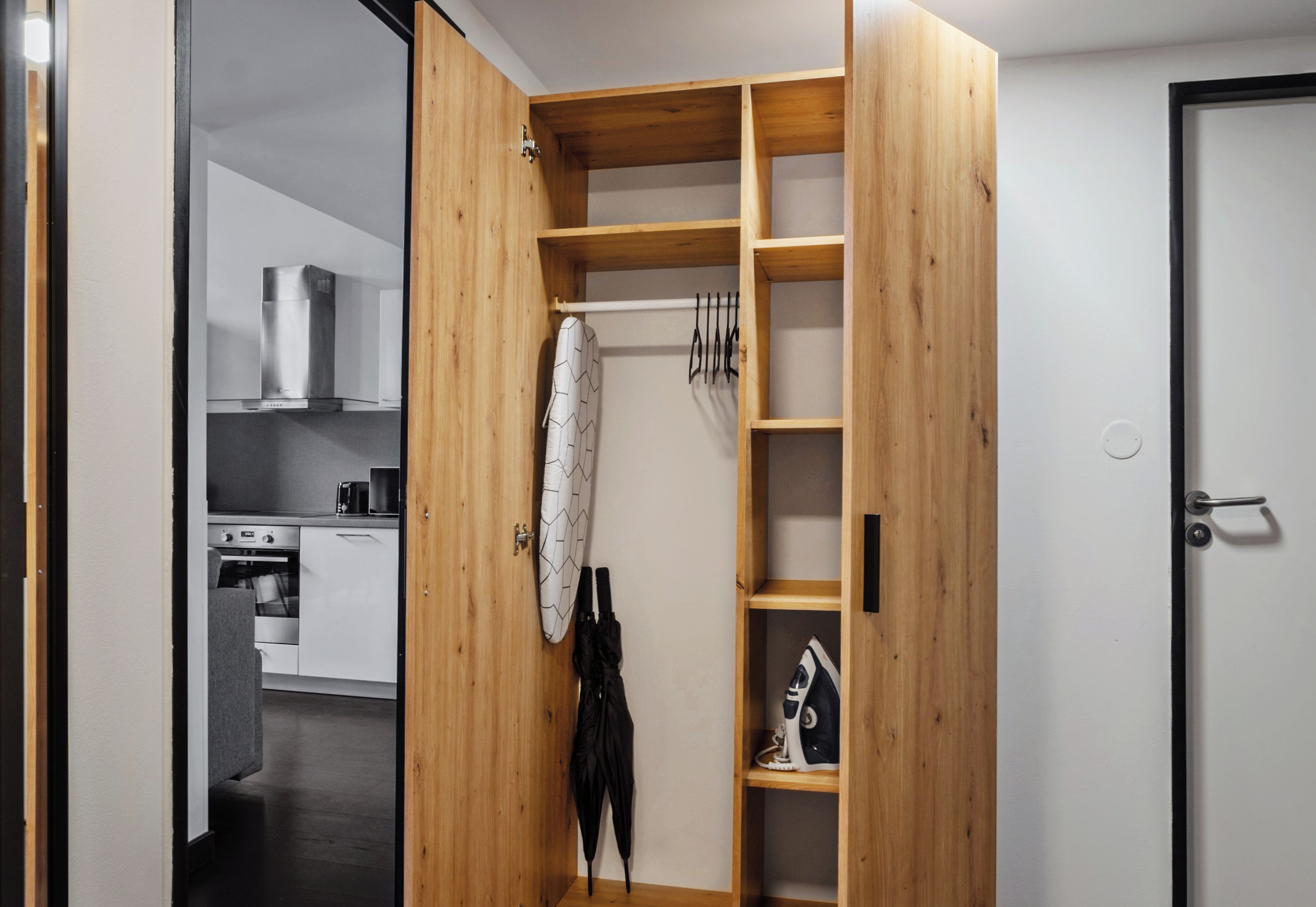 A compact kitchen with modern appliances, a wooden wardrobe with shelves, and a door leading to another room.