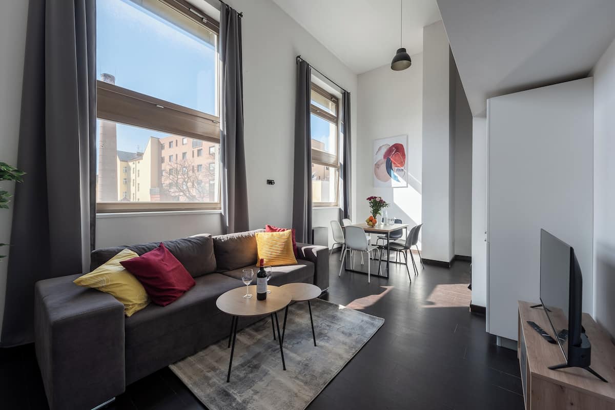 Een moderne woonkamer met een grijze bank, een klein rond tafeltje met een wijnglas, en een tv op een houten dressoir.
