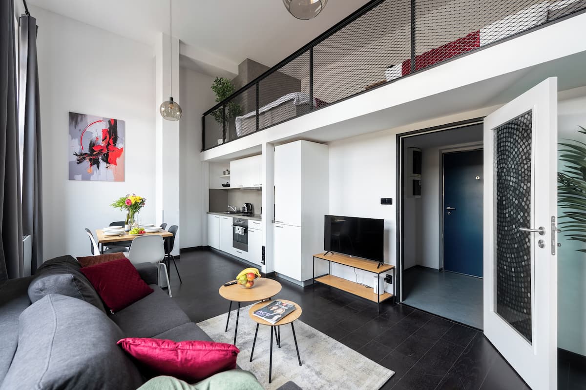 Een moderne woonruimte met een grijze bank, een klein rond tafeltje met een boek, een televisie op een meubelstuk en een keuken in de achtergrond.