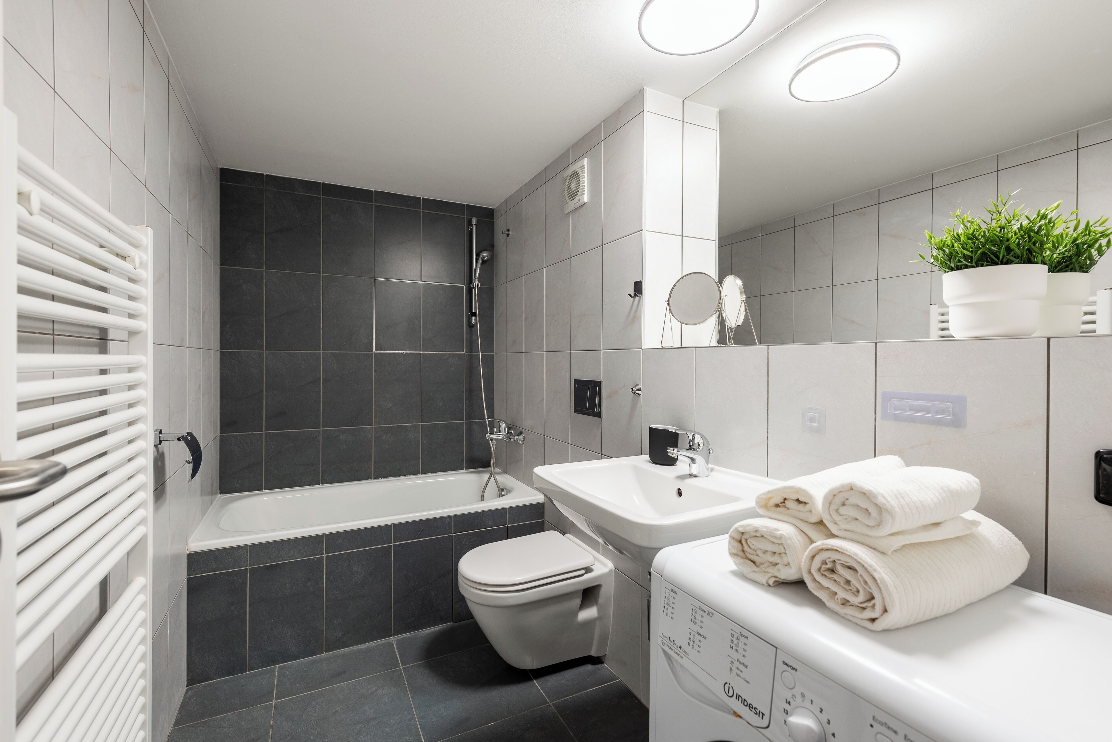 Een moderne badkamer met een bad, toilet en wastafel. Handdoeken liggen op de wastafel gestapeld en er staat een potplant op het aanrecht.
