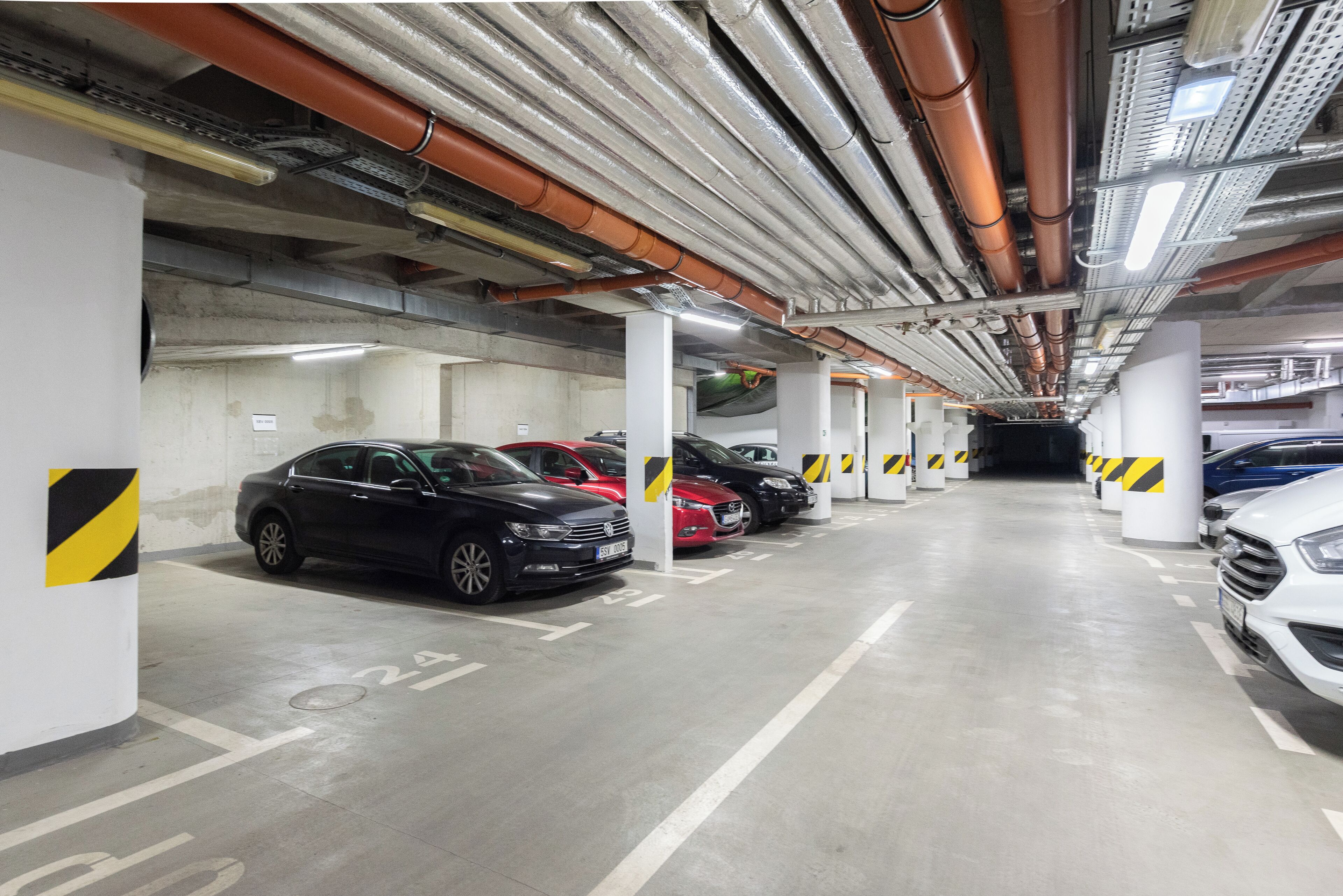 En parkeringskælder med flere rækker af parkerede biler, betonstolper og ovenlys.