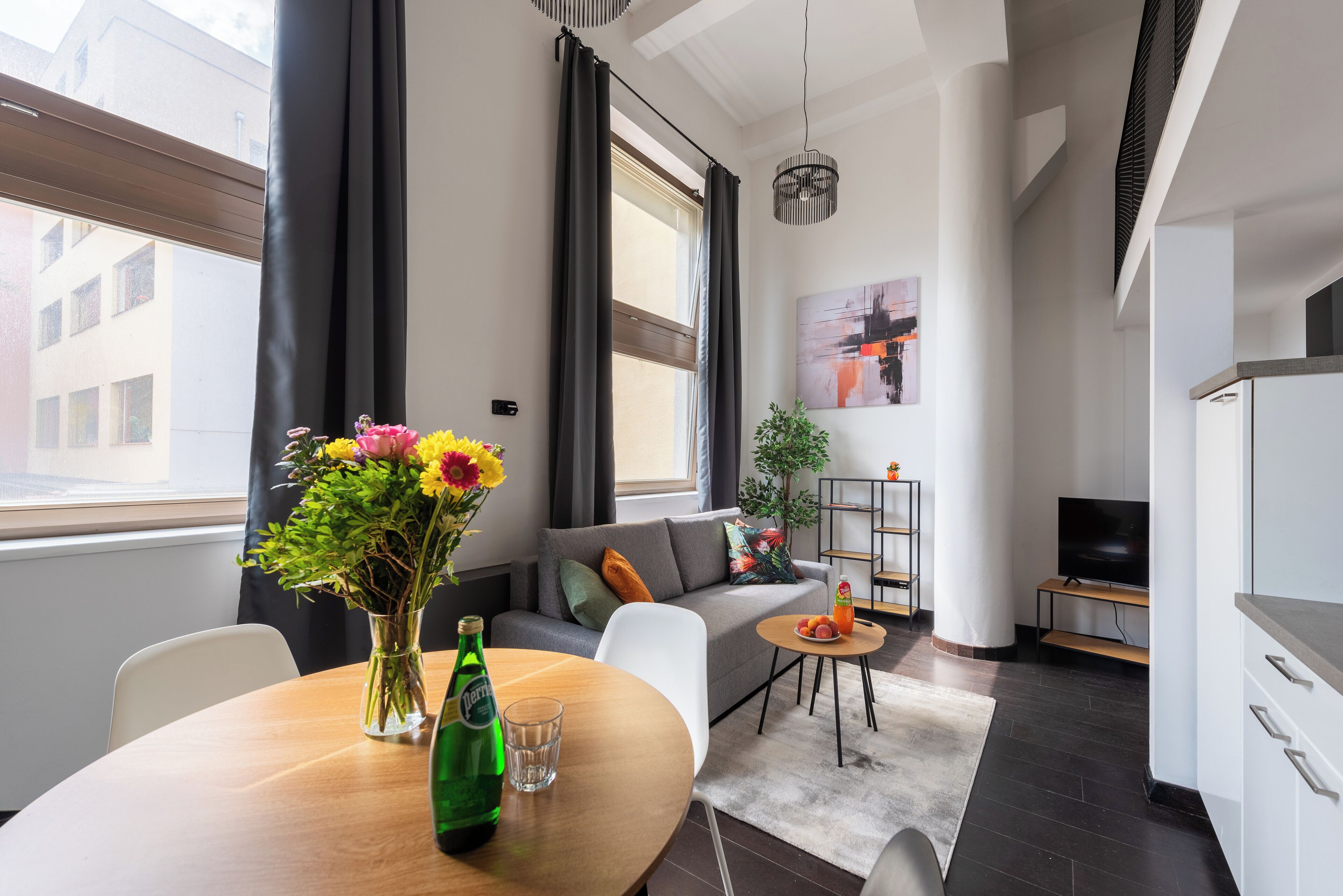 Ein modernes Wohnzimmer mit einer Couch, einem Esstisch mit Blumenvase, einem Fernseher und einer Küchenzeile.