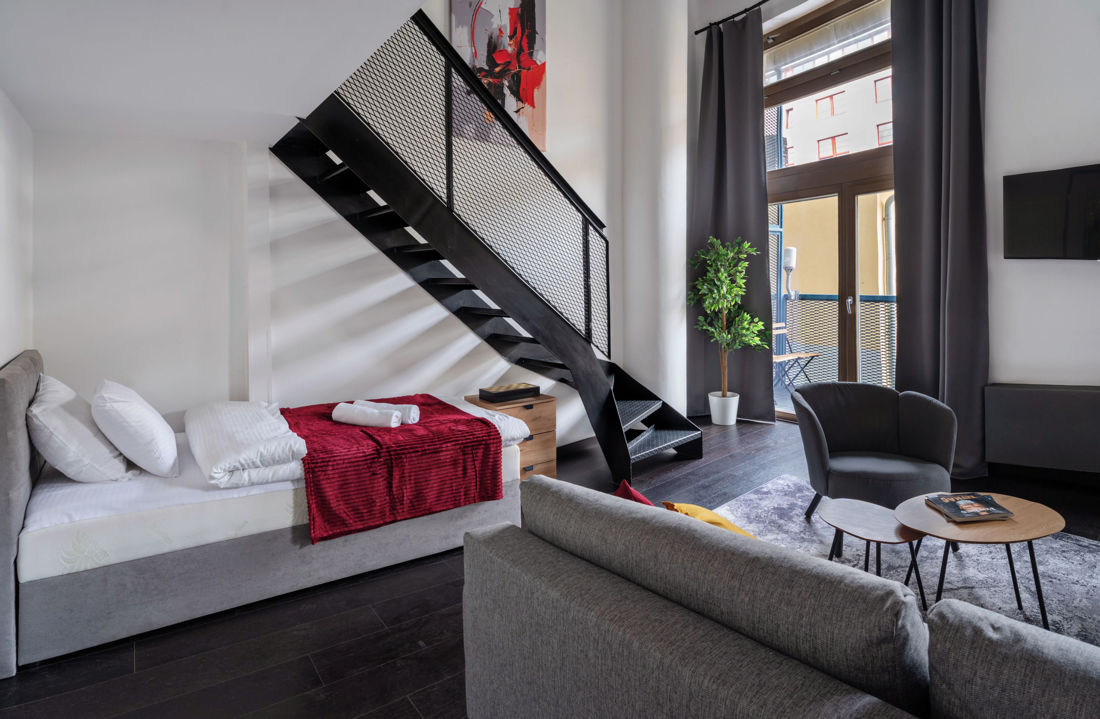 A modern living room with a grey sofa, a red throw blanket, a black staircase, a grey armchair, and a small round table with a book.