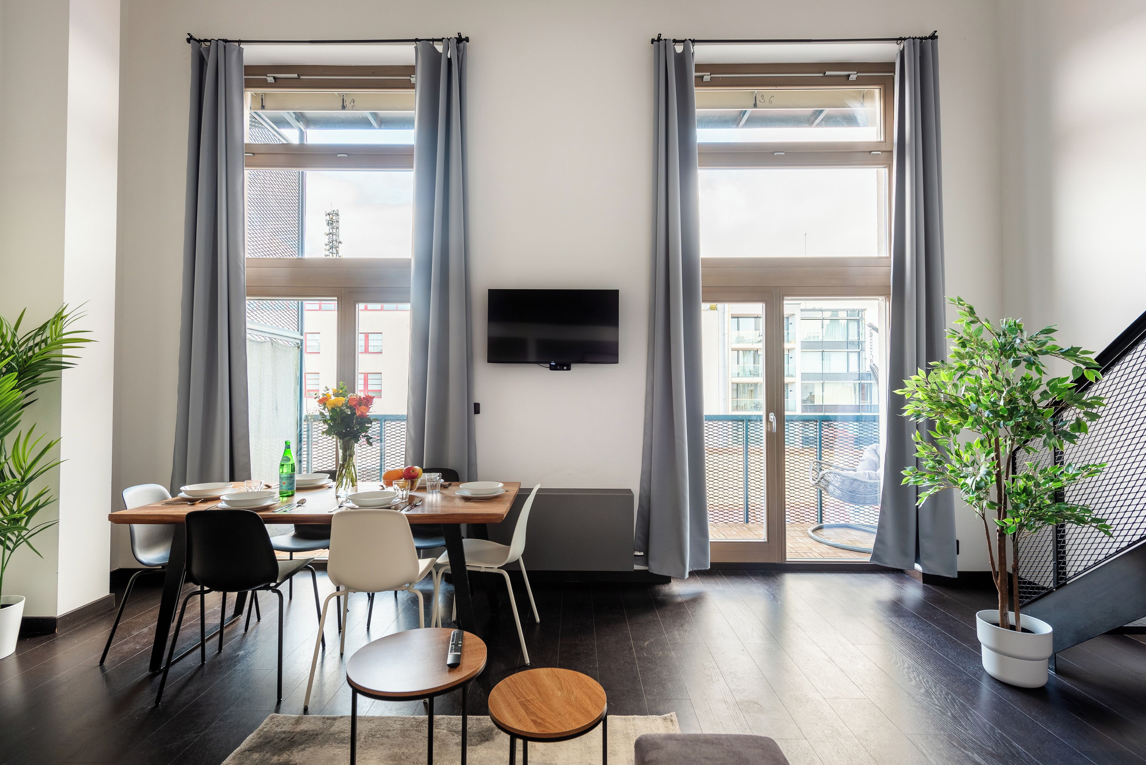 Un espace repas moderne avec une table dressée pour deux personnes, un téléviseur fixé au mur et de grandes fenêtres avec des rideaux.