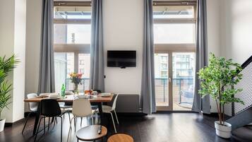 Un espace repas moderne avec une table dressée pour deux personnes, un téléviseur fixé au mur et de grandes fenêtres avec des rideaux.