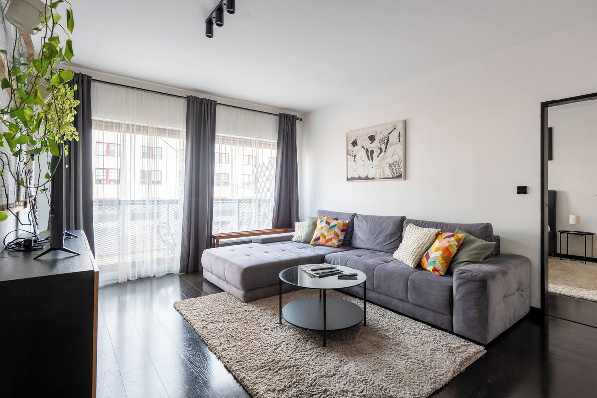 Een moderne woonkamer met een grijze bank, een ronde salontafel en een groot raam met gordijnen.
