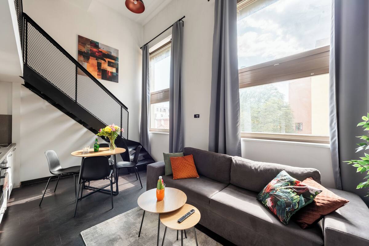 Een moderne woonkamer met een bank, een eettafel met stoelen, een trap en grote ramen met gordijnen.