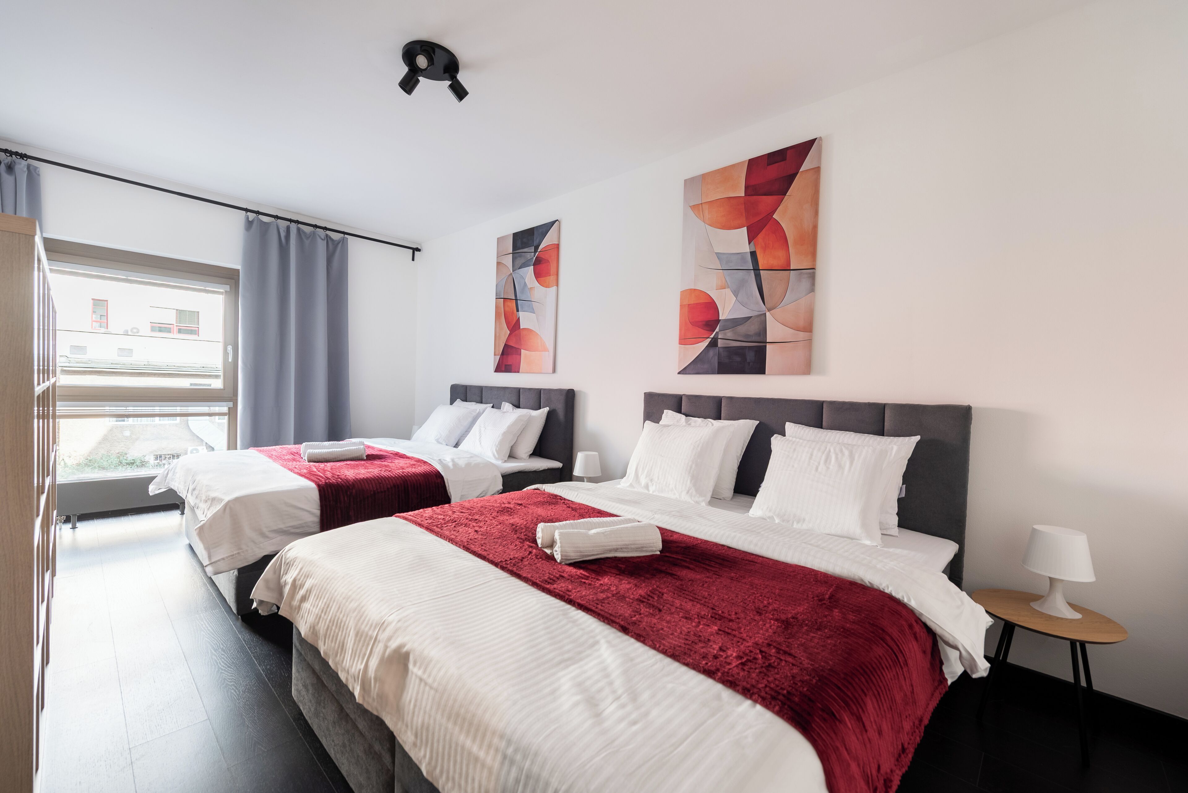 Habitación de hotel con dos camas, una ventana con cortinas, un cuadro abstracto en la pared y una mesita con lámpara.