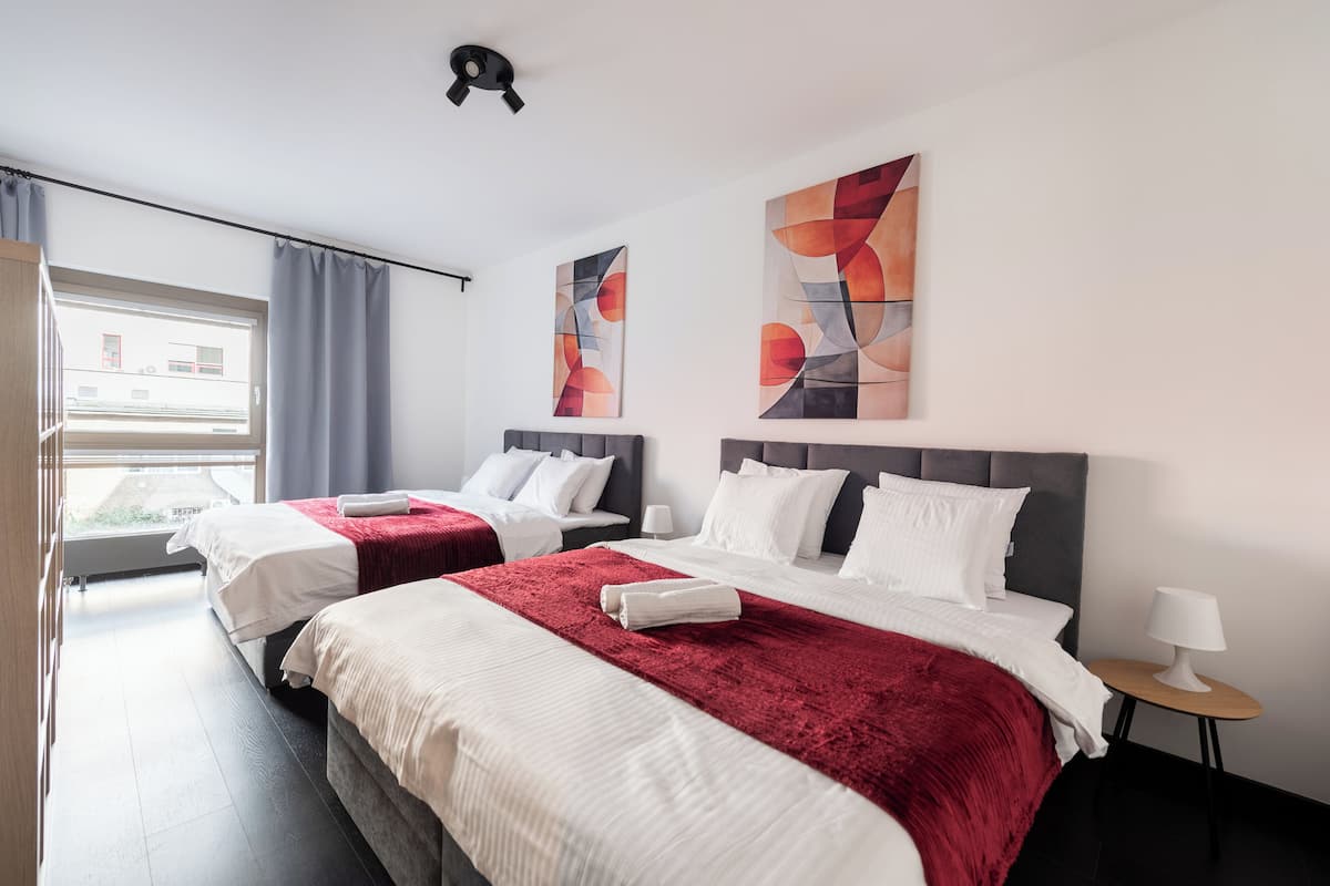 Een hotelkamer met twee bedden, een raam met gordijnen, abstracte wanddecoratie en een tafeltje met een lamp.