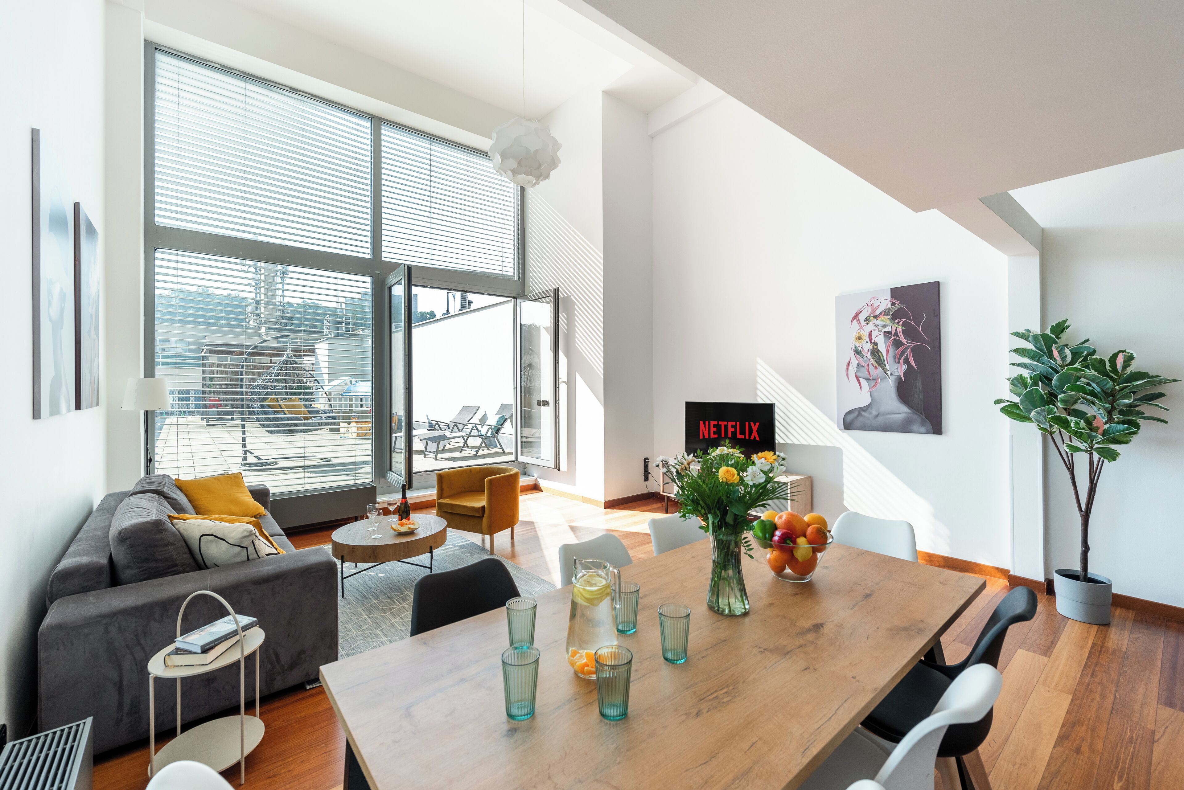 A modern living room with a sofa, coffee table, and dining table. There is a large window with blinds, a plant, and a sign that says "NETFLIX".