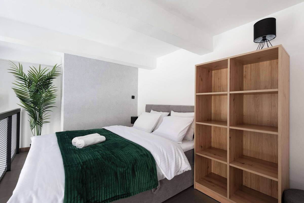 Een moderne slaapkamer met een bed, een groene deken, witte kussens, een groot houten boekenrek en een potplant.