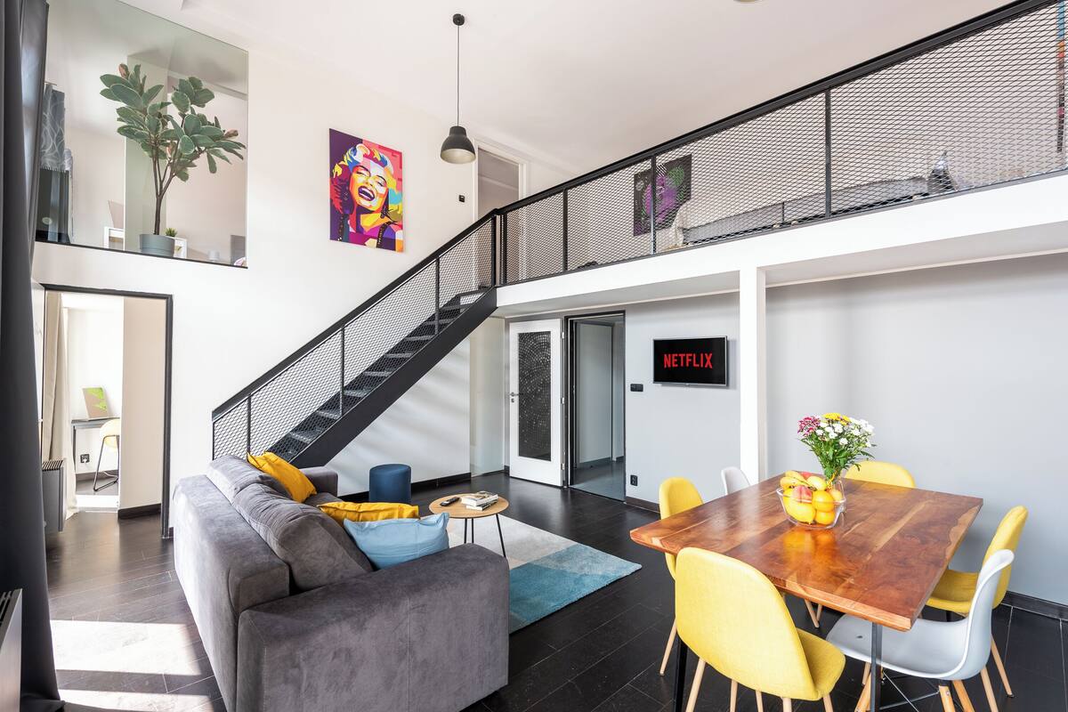 Een moderne woonruimte met een grijze bank, gele stoelen, een houten eettafel en een trap met een gaasreling.