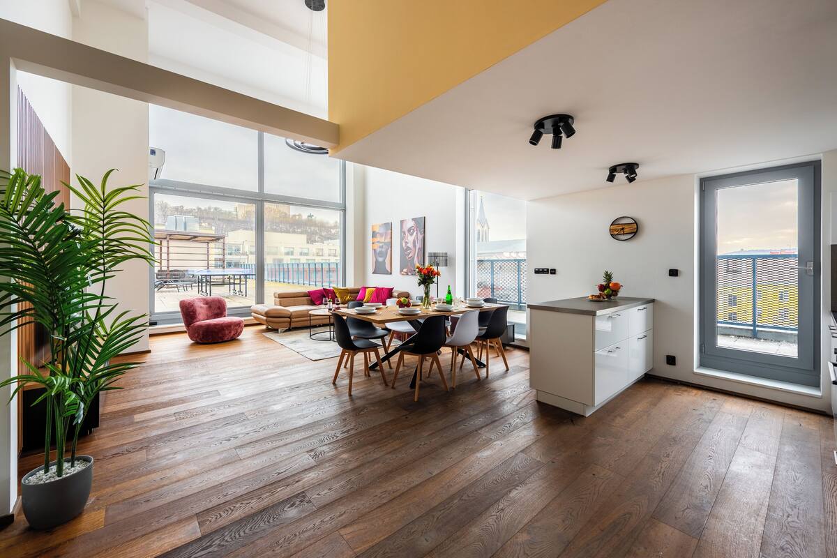Een ruime woonkamer met een houten vloer, een eettafel met stoelen, een rode fauteuil, een potplant en een stadszicht door grote ramen.