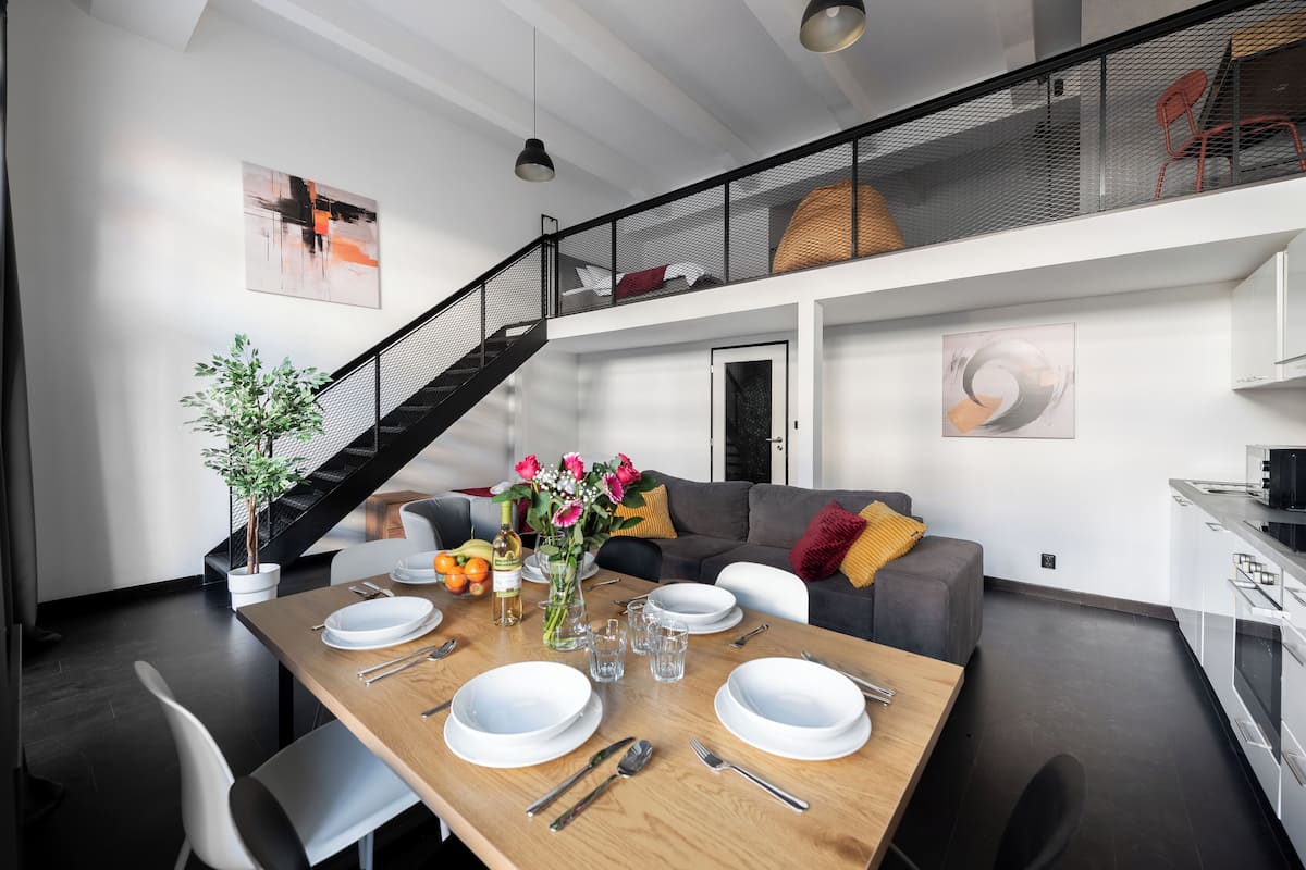 Een moderne eetruimte met een houten tafel voor vier personen, een trap en een zithoek met een grijze bank en een rode fauteuil.