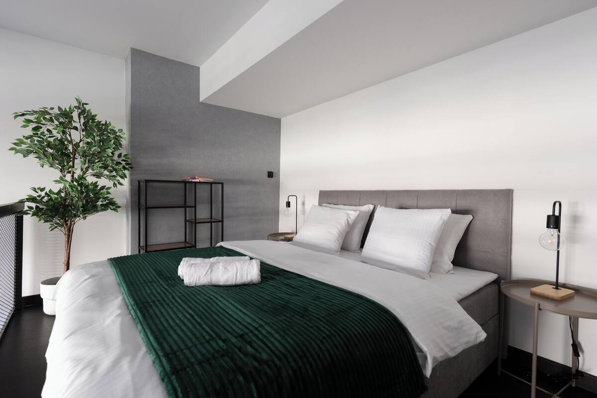 Een moderne hotelkamer met een groot bed, een groene plaid, een nachtkastje met een lamp en een potplant.