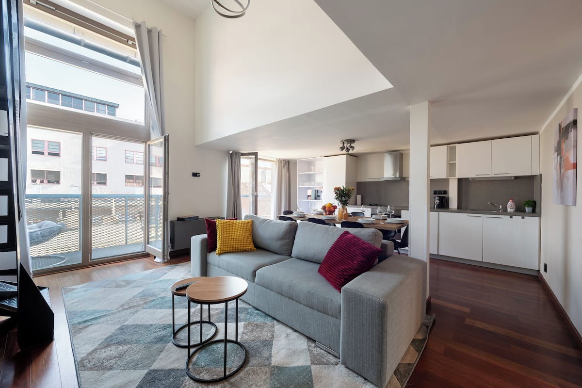 Een modern appartement met een grijze bank, een klein rond tafeltje en een keuken op de achtergrond.