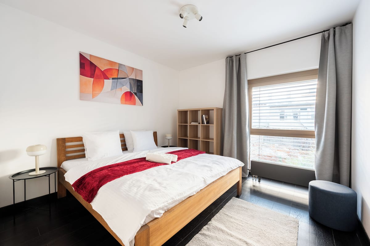 Een moderne slaapkamer met een houten bed, een rode plaid, een wit kussen, een nachtkastje met een lamp, een boekenkast en een groot raam met gordijnen.