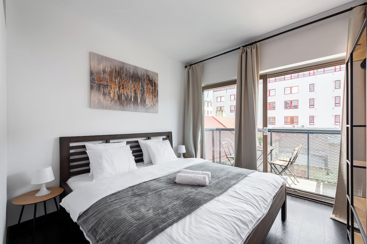 Een moderne hotelkamer met een groot bed, een klein nachtkastje, een schilderij aan de muur, en een balkon met uitzicht op gebouwen.