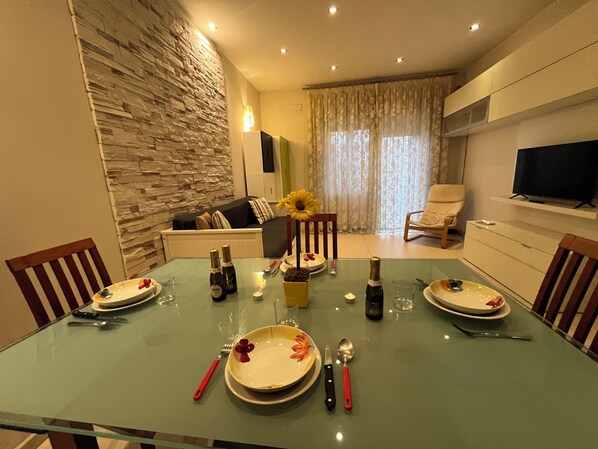 A dining area with a glass table, plates, cutlery, and a bottle of champagne. There is a sofa and a TV in the background.
