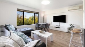 A modern living room with a sofa, a coffee table, a dining table with chairs, a flat-screen TV mounted on the wall, and a large window with a view of trees.