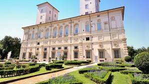 Un grande edificio storico dall'architettura simmetrica, con un cortile centrale affiancato da giardini curati e una statua.