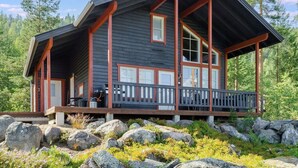 Une cabane dotée d’une véranda, de grandes fenêtres et d’un toit en pente, entourée de rochers et d’arbres.