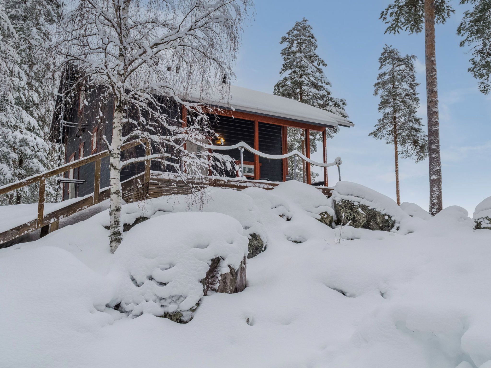 Una cabaña de madera cubierta de nieve con techo rojo, rodeada de árboles y rocas.