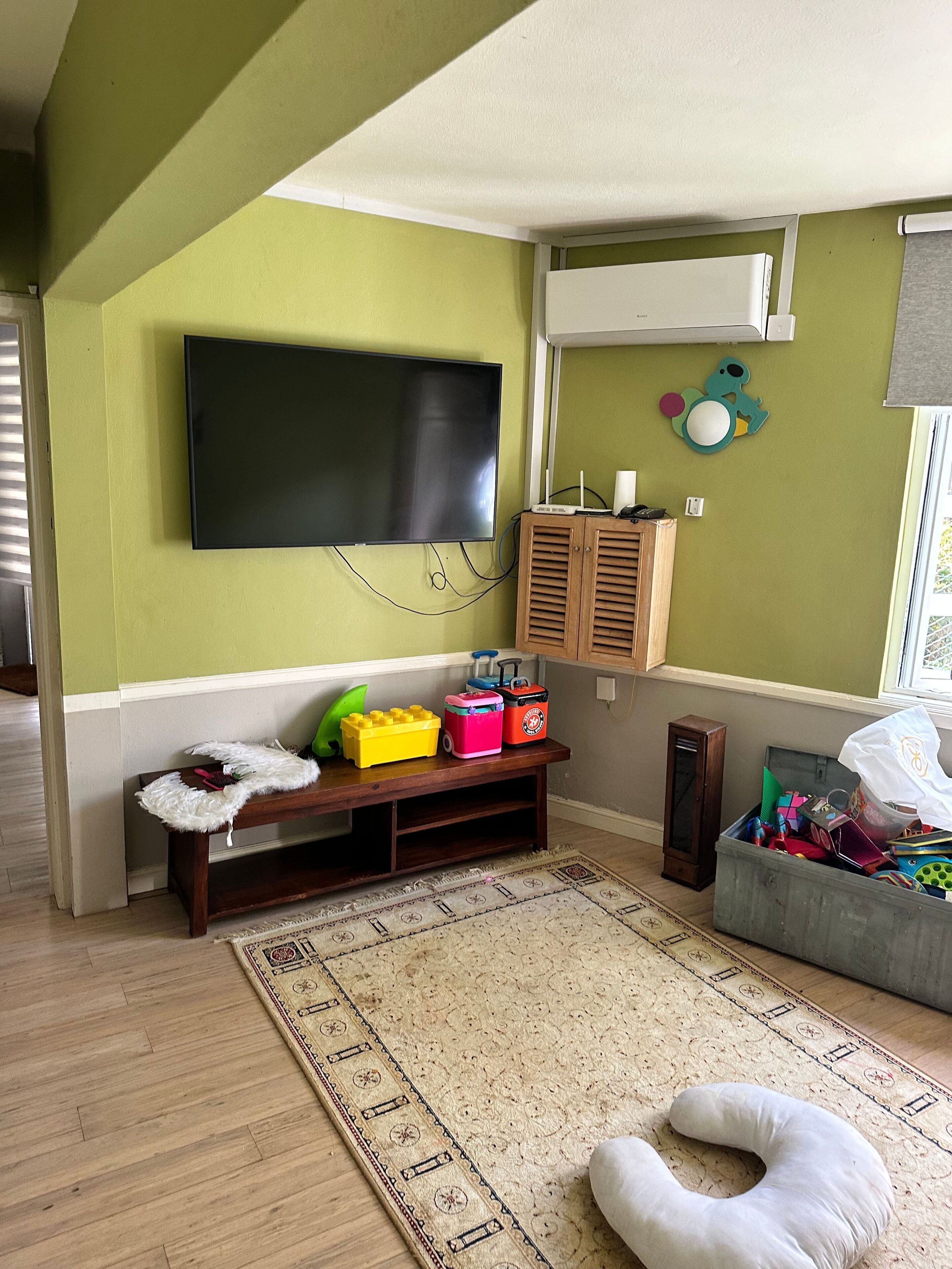A living room with a flat-screen TV mounted on the wall, a wooden cabinet, a colorful play area with toys, and a patterned rug on the floor.