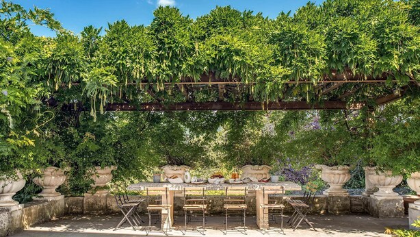 Een buitenruimte met een pergola begroeid met groen, stenen plantenbakken en een stenen tafel met stoelen.
