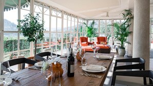 Een eetgedeelte met een houten tafel gedekt voor een maaltijd, grote ramen met uitzicht op de omgeving, en een zithoek met oranje stoelen.