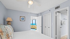 A bedroom with a bed, a nightstand, a lamp, a framed picture, and a view of the ocean through an open door.