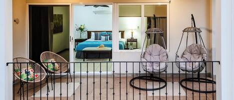 A balcony with wicker chairs, a hanging chair, and a bed visible through a glass door.